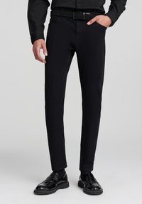 Czarne spodnie slim fit wykonane z tkaniny bawełnianej, charakteryzujące się gładką strukturą, zamkiem błyskawicznym oraz kieszenią z przodu. Noszone z błyszczącymi czarnymi butami.