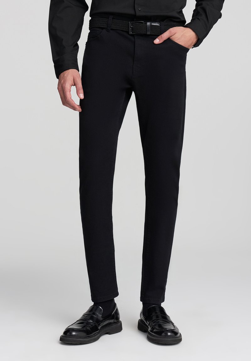 Czarne spodnie slim fit wykonane z tkaniny bawełnianej, charakteryzujące się gładką strukturą, zamkiem błyskawicznym oraz kieszenią z przodu. Noszone z błyszczącymi czarnymi butami.