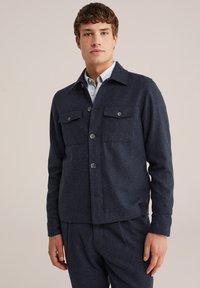 Marineblauwe wolmix jas met knoopsluiting, twee borstzakken en een kraag. Soepele textuur, relaxte pasvorm en minimale details.