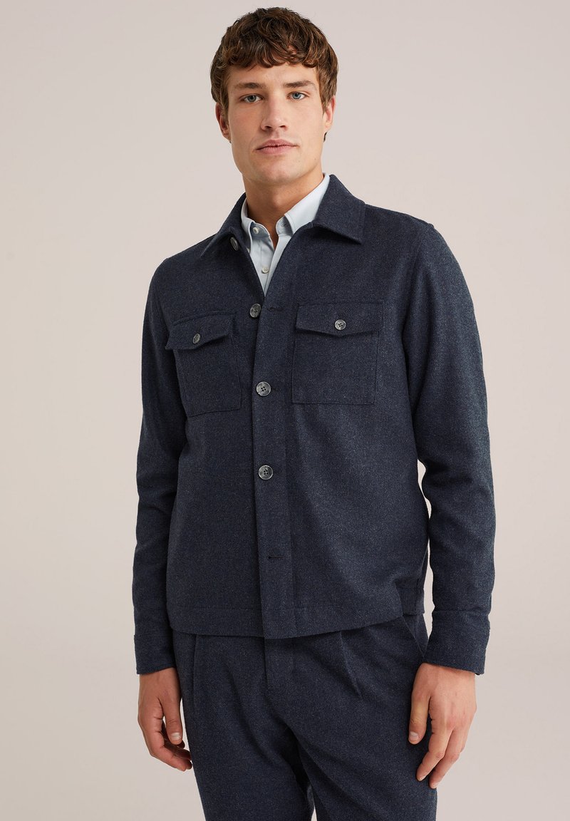 Marineblauwe wolmix jas met knoopsluiting, twee borstzakken en een kraag. Soepele textuur, relaxte pasvorm en minimale details.