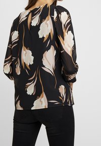 Blusa nera con motivo floreale in crema e rosa pallido. Presenta una scollatura a forma di occhiello sul retro e maniche a sbuffo. Realizzata in tessuto leggero.