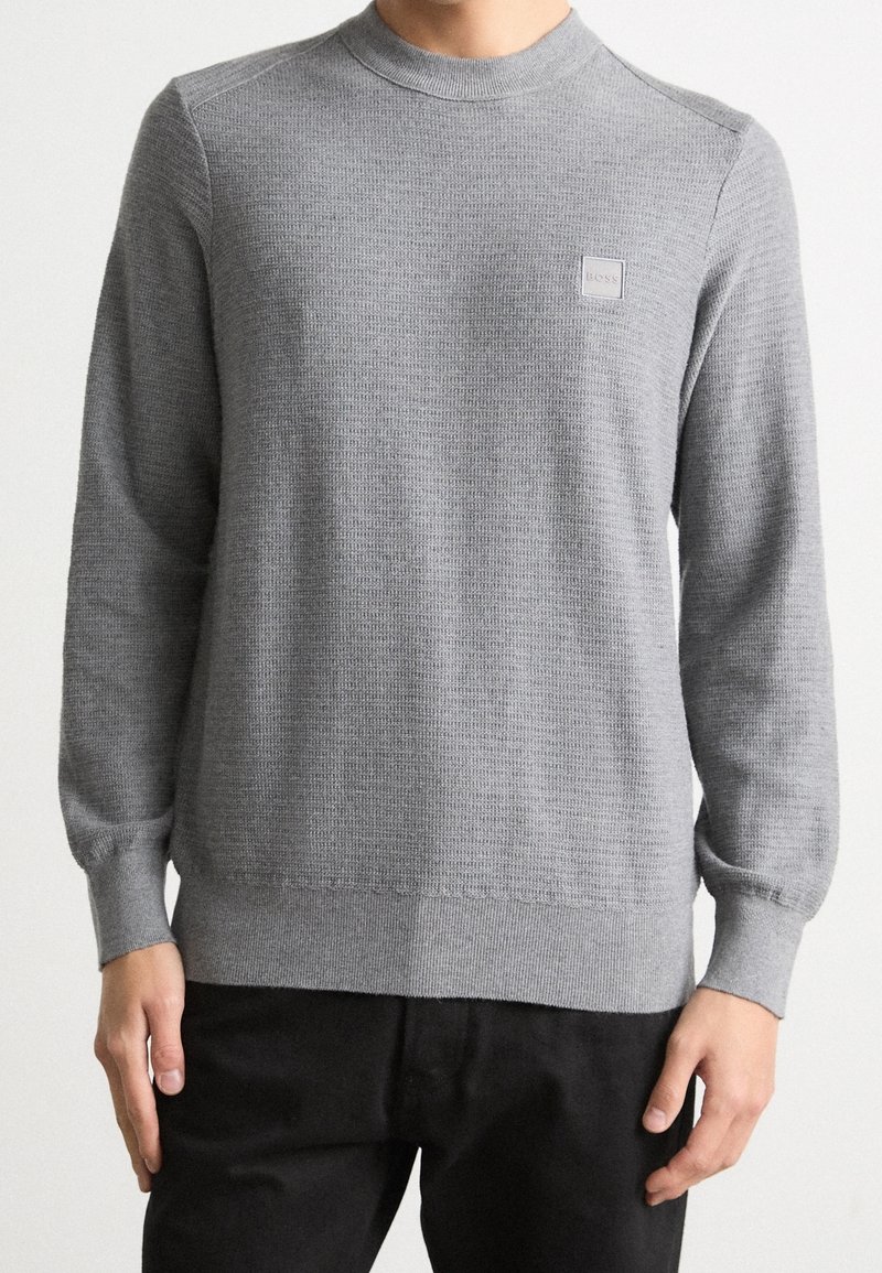 Homme portant un pull gris clair texturé à manches longues et col rond avec un petit logo carré Boss sur la poitrine, associé à un pantalon noir.