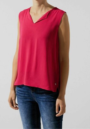 Bezramienna czerwona bluzka z zagiętym kołnierzykiem w kształcie litery V, noszona z niebieskimi dżinsami przez osobę stojącą na jasnym tle.