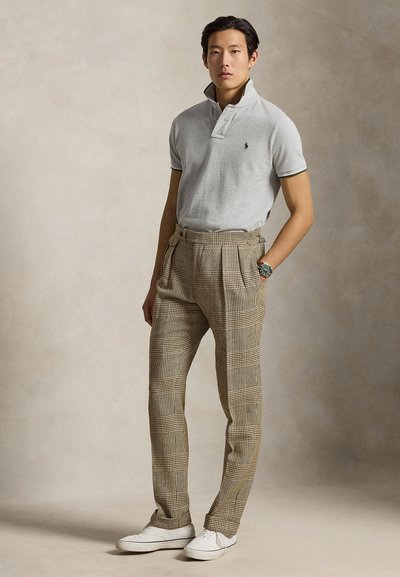 Homme debout portant un polo gris clair, un pantalon beige à carreaux taille haute, des baskets blanches et une montre au poignet gauche sur fond uni.