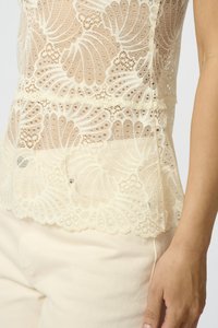 Lace toppi vaaleassa kermassa, jossa on monimutkaisia floristikuvioita, aaltoileva helma ja läpikuultava rakenne. Yhdistetty vaalean kermanvärisiin kangashousuihin.