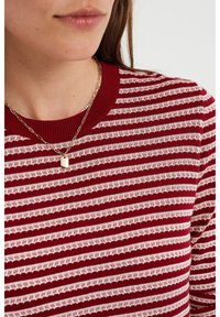 Punainen neulepusero, jossa on valkoisia vaakaraitoja, teksturoitu kuvio ja resorimainen pääntie. Kultainen riipuskaulakoru suorakulmaisella koristeella.