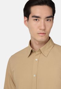 Béžová košile na knoflíky vyrobená z měkkého materiálu, s klasickým límečkem, předním zapínáním a bílými knoflíky. Hladká struktura a přiléhavý střih.