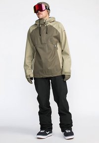 Olive green and beige jacket with a half-zip design, paired with black pants and snow boots. Accessories include a beanie and reflective goggles.