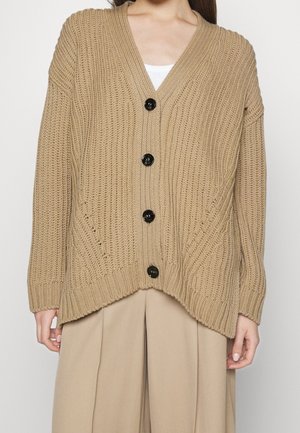 Femme portant un cardigan en tricot épais beige oversize avec des boutons noirs et un pantalon large beige, visible du cou jusqu'à mi-cuisse.
