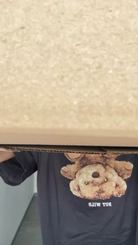 Caixa de cartão castanha segurada com duas mãos. O topo da caixa tem uma textura suave, isenta de marcas. A camisa escura apresenta um gráfico de um ursinho de pelúcia.