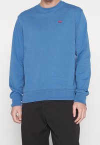 Sudadera azul hecha de tejido suave, con un cuello redondo y puños acanalados. Incluye un pequeño logo rojo de Levi's en el pecho.