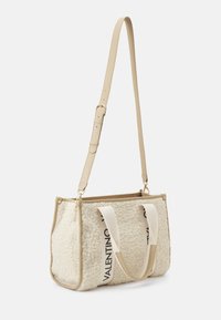 Béžová kabelka z ovčí vlny s koženými detaily. Má nastavitelný ramenní popruh a dvě horní uchy. Na přední straně výrazné logo.