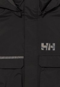 Schwarze Jacke mit Reißverschluss und Klappentaschen, ausgestattet mit reflektierendem "HH"-Logo und horizontalem reflektierendem Streifen auf der linken Tasche.
