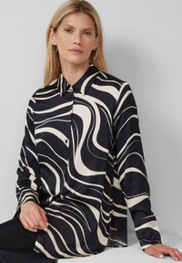 Donna con lunghi capelli biondi, che indossa grandi orecchini a cerchio e una camicia a bottoni con un motivo astratto in bianco e nero, seduta su uno sfondo grigio.