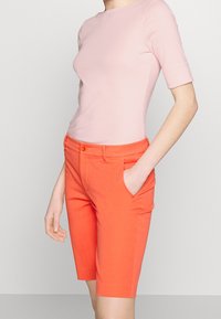 Mujer vistiendo una blusa ajustada de color rosa claro y pantalones cortos corales brillantes hasta la rodilla con bolsillos, de pie frente a un fondo liso y claro.