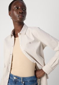 Light beige button-up shirt with a soft texture, featuring a small embroidered design. Paired with a fitted beige top and blue denim jeans.
