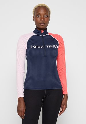 Frau mit kurzen blonden Haaren, die ein marineblaues, geripptes Oberteil mit pinken und korallenfarbenen langen Ärmeln und einem Frontreißverschluss trägt, auf dem das Logo "KARI TRAA" zu sehen ist.