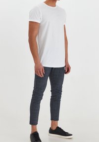 Homme portant un t-shirt blanc uni à manches courtes, un pantalon slim à carreaux gris et des baskets noires à semelles blanches, debout sur un fond blanc.