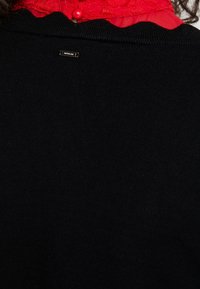 Schwarzer Pullover mit geripptem Kragen und gewelltem Rand. Verfügt über ein kleines metallisches Etikett auf der Rückseite. Darunter befindet sich eine rote Spitzen Detail.