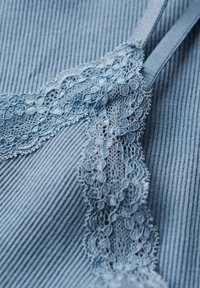 Lichtblauwe stof met een geribbelde textuur, voorzien van delicate kantdetails langs de rand en dunne bandjes. Een subtiele glans accentueert het materiaal.