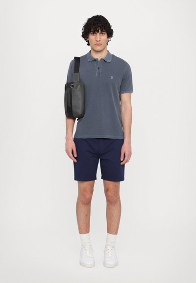 Polo shirt blu scuro, pantaloni corti navy e sneaker bianche. Una borsa a spalla grigia con una tracolla poggia su una spalla. Sfondo semplice.