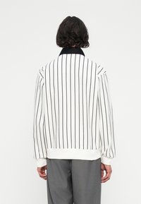 Bílý pruhovaný pulovr s vertikálními černými pruhy. Měkká látka, žebrované manžety a černý límec. Jednoduchý, minimalistický design.