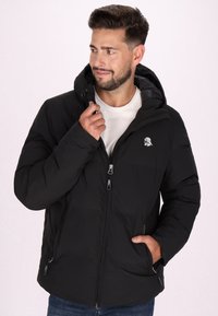 Giacca a vento nera con cappuccio, dotata di zip frontale, tasche laterali e un logo sottile sul petto. Materiale liscio e texturizzato.
