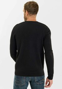 Uomo che indossa un maglione lungo nero e jeans blu, rivolto di spalle contro uno sfondo chiaro e uniforme.