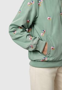 Zielona kurtka typu bomber z haftowanymi różowymi, żółtymi i niebieskimi kwiatami, noszona z ręką w bocznej kieszeni i widocznymi beżowymi spodniami.