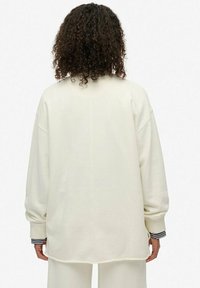 Sudadera de color crema, de corte oversize, con hombros caídos y puños de canalé. Presenta una textura suave y un dobladillo redondeado. Detalles a rayas en los puños.