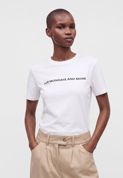 Femme aux cheveux courts portant un t-shirt blanc avec le texte "FOR MONDAYS AND MORE" et un pantalon beige taille haute, mains dans les poches, fond neutre.