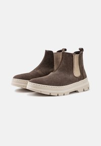 Botas de tobillo de ante marrón con paneles laterales elásticos y una suela de goma blanca gruesa. Presentan una punta redondeada y lengüetas para facilitar su puesta.