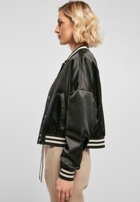 Veste bomber en satin noir avec des poignets et un ourlet côtelés à rayures blanches et noires, dotée de boutons-pression et de poches latérales pour une touche supplémentaire.