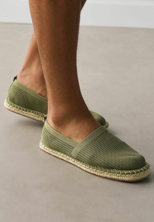 Legs wearing olive green mesh espadrilles with woven jute soles, standing on a smooth gray floor.