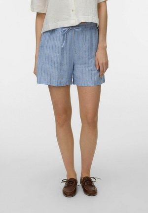 Kobieta w niebiesko-białych spodenkach w pionowe paski z troczkiem, białej bluzce z krótkim rękawem i brązowych butach na sznurowadła, stojąca na białym tle.