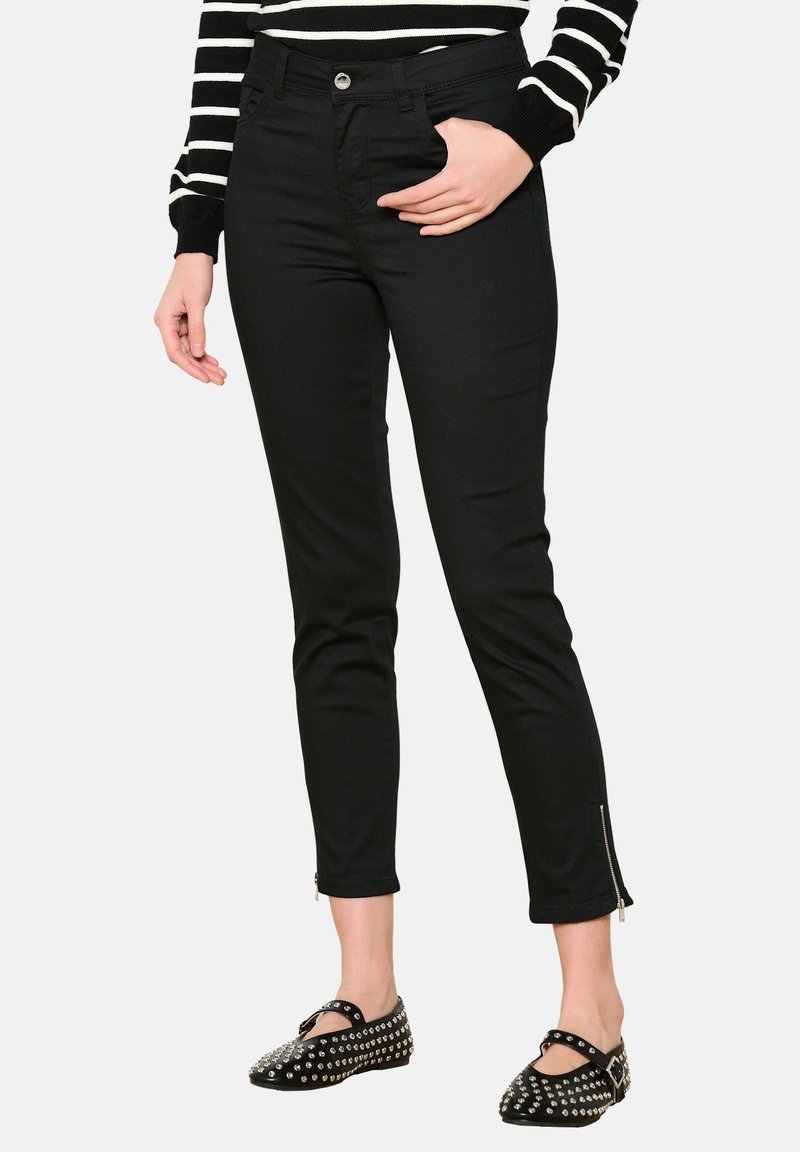 Pantalones skinny de mezclilla negra con una cintura alta, bolsillos frontales y detalles de cremallera en el tobillo, combinados con una blusa a rayas y zapatos con tachuelas.