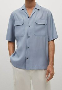 Chemise à manches courtes bleu clair en tissu lisse avec deux poches poitrine et boutons noirs, présentant une coupe décontractée et un col de type camp.