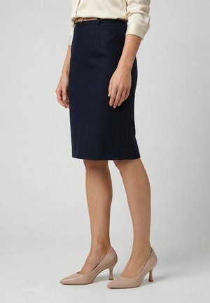 Kvinde iført en knælang navy blyantsnederdel, beige bluse med opkørte ærmer og beige midthælspidsede sko, stående sideværts.