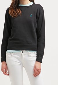 Svart sweatshirt med rundad halsringning, långa ärmar och en turkos logotyp på bröstet, i kombination med vita jeans. Slät textur, avslappnad passform.