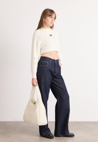 Woman wearing a cropped white sweater and dark wide-leg jeans, holding a large beige handbag and standing on a gray floor.