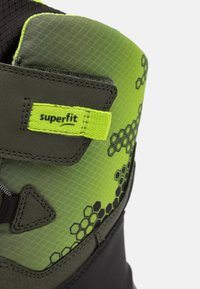 Zelená a čierna čižma s hexagonálnym vzorom, texturovaný povrch a štítok "superfit". Vyznačuje sa pevnou konštrukciou a vzorovanými akcentmi.