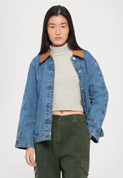 Τζάκετ από τζην με καφέ γιακά, κλείσιμο με κουμπιά και μεγάλες τσέπες; φοριέται επάνω από μια ριγέ τσιχλική μπλούζα και συνδυάζεται με πράσινα cargo παντελόνια.