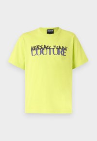 Neon žluté bavlněné tričko s krátkými rukávy. Obsahuje nápis "VERSACE JEANS COUTURE" v tučném, stylizovaném fialovém grafickém písmu.