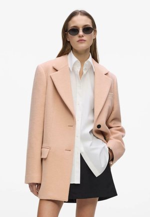 Vrouw met zwarte ovale zonnebril, oversized beige jas, wit overhemd en zwarte rok, staand tegen een effen witte achtergrond.