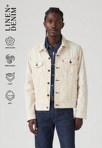 Linnenmix, licht beige jacket met knoopsluiting, borstzakken en contrasterende stiksels, gedragen over een donkerdenim overhemd en blauwe spijkerbroek.