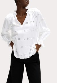 Blusa de algodón blanca con bordados de ojal, cuello fruncido, mangas largas y puños en forma de ondas, combinada con pantalones negros.