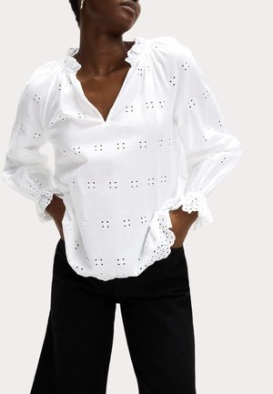 White cotton blouse with eyelet embroidery, frilled neckline, long sleeves, and scalloped cuffs, paired with black pants.