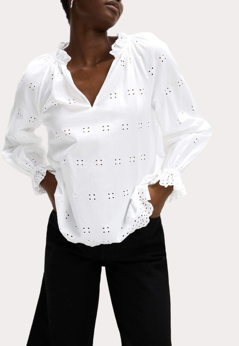 Blusa de algodón blanca con bordados de ojal, cuello fruncido, mangas largas y puños en forma de ondas, combinada con pantalones negros.