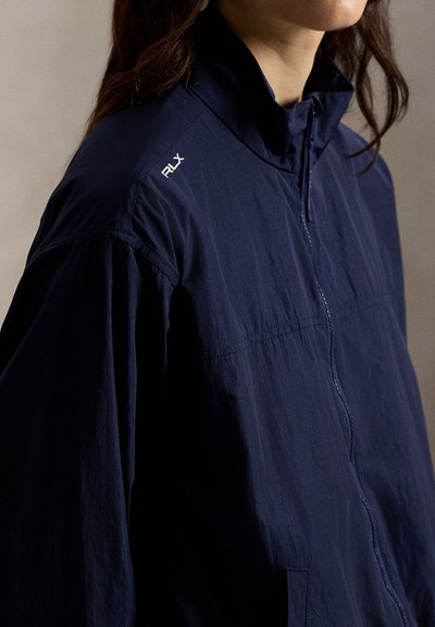 Marineblå, lett jakke med glidelås foran, ståkrage, teksturert stoff og hvit "RLX"-logo på venstre skulder. Reflekterende detaljer.