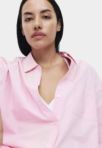 Chemise surdimensionnée rose pâle en tissu doux, avec un devant à boutons et une poche poitrine. Motif rayé texturé visible.